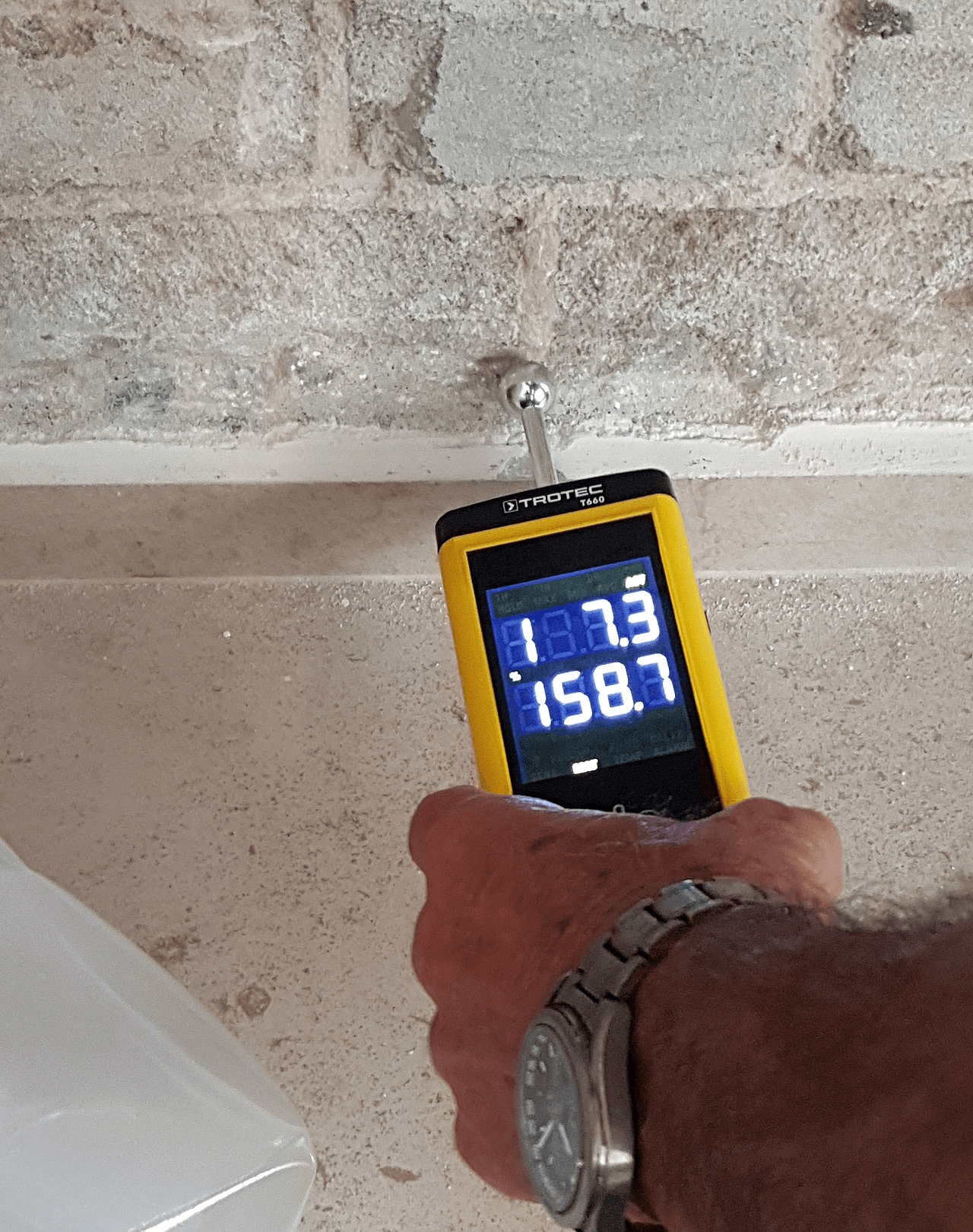 Handheld moisture meter held against a kitchen wall showing an elevated reading on its display